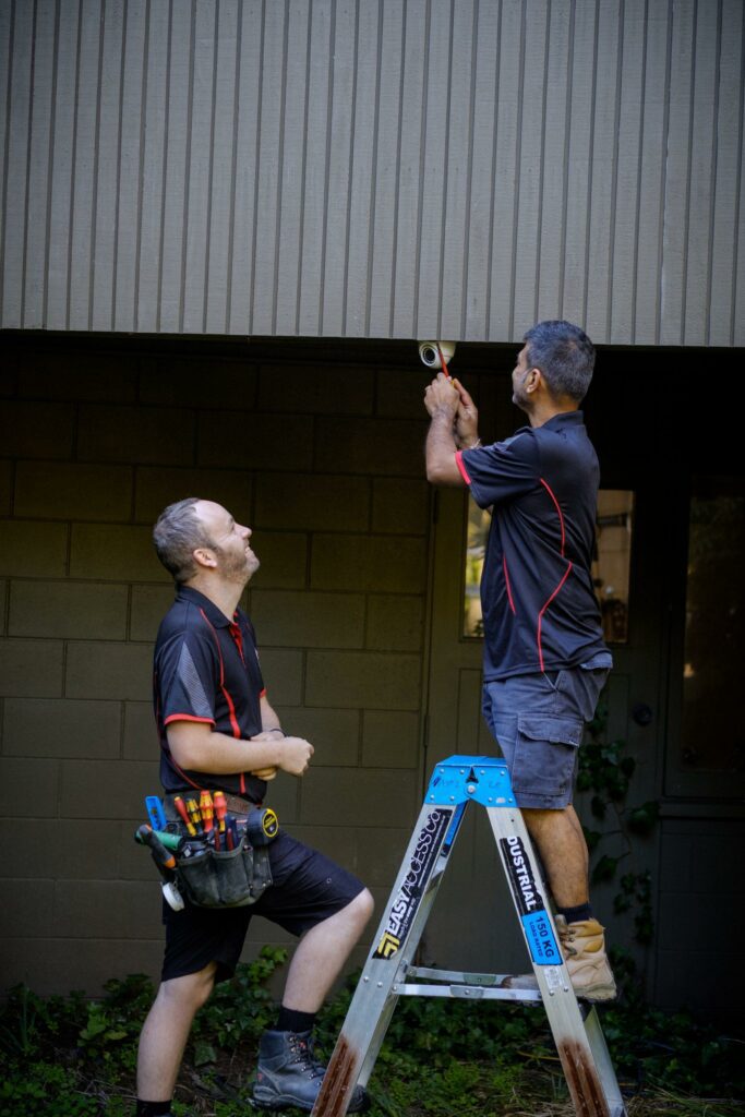 Two 2E Electrical technicians installing outdoor equipment with one on a ladder and one assisting